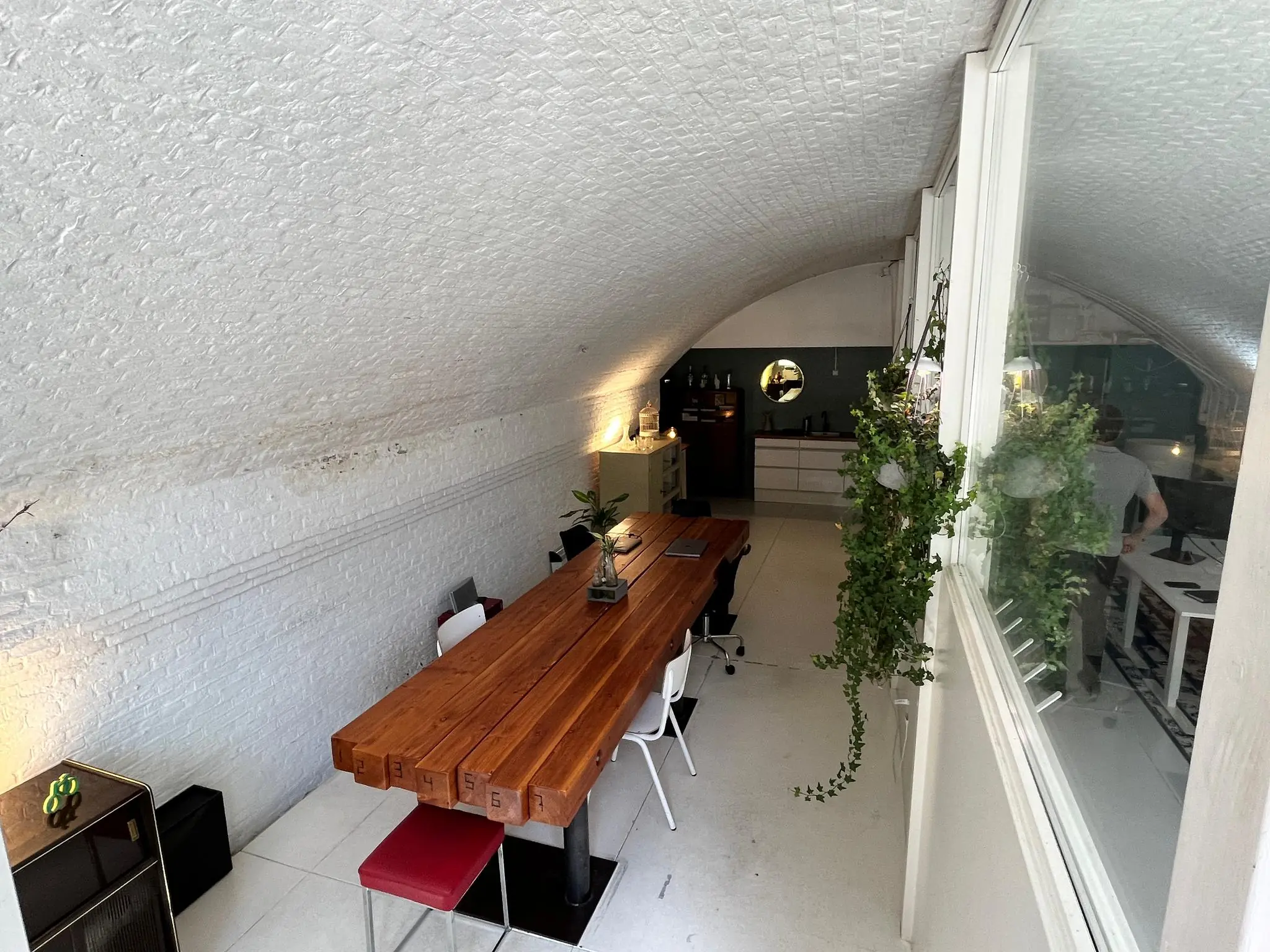 Vergaderruimte onder een witgewelfd plafond met een lange houten tafel, stoelen en uitzicht op een aangrenzend kantoor via een raam.